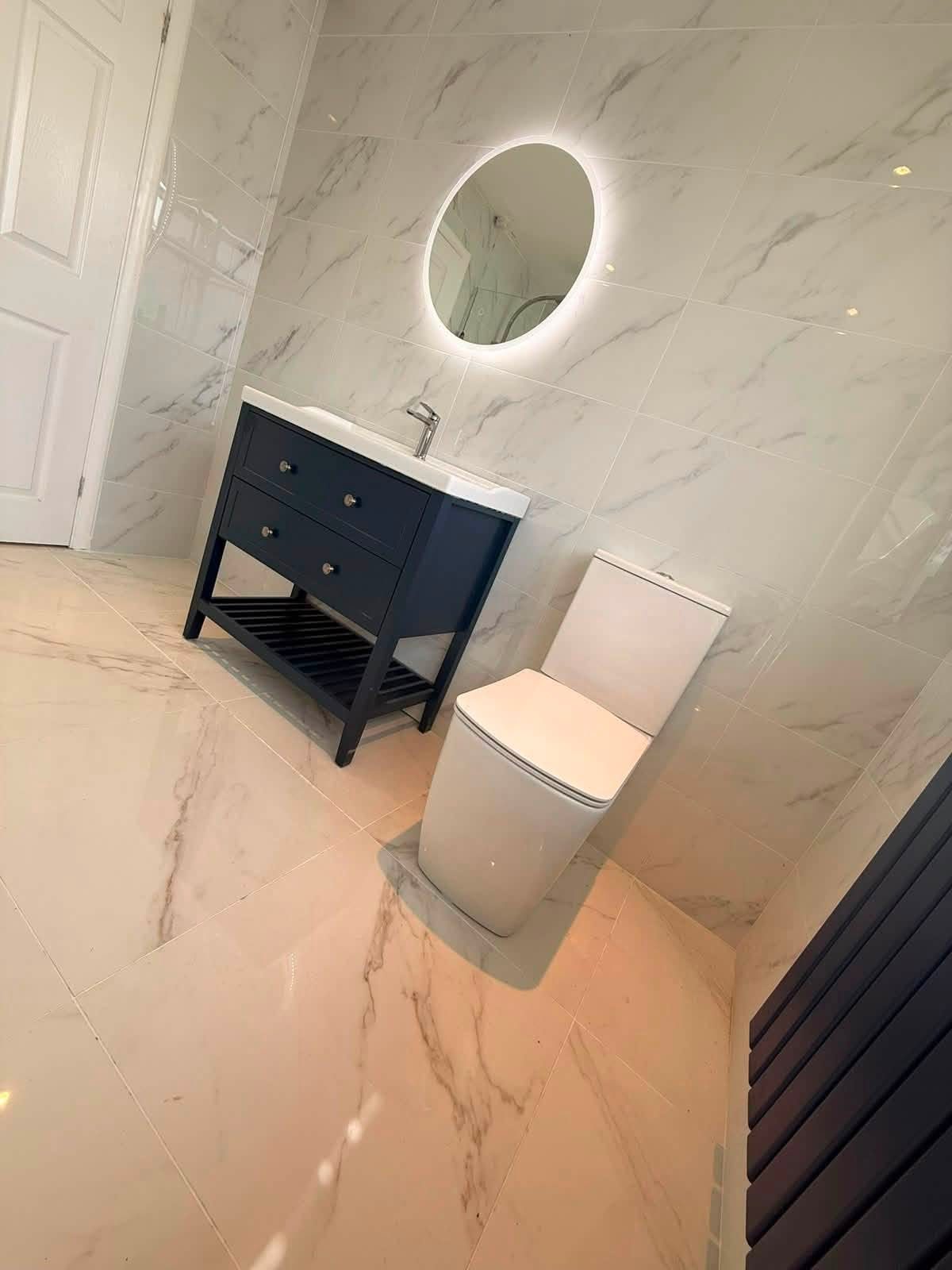 Completed bathroom with dark vanity unit, illuminated round mirror and marble-effect tiling